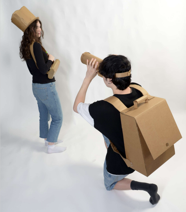 woman wearing cardboard hat and purse while man kneels down wearing a cardboard backpack and using a cardboard camera