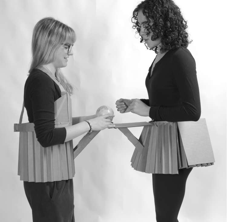 two women wear cardboard skirts that have a ledge on the front of them that connect to form a table. A globe balances on the table between them. 