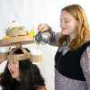 a woman wears a cardboard headdress that has a plate and cups attached to it while another woman pours coffee into one of the cups