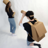 woman wearing cardboard hat and purse while man kneels down wearing a cardboard backpack and using a cardboard camera