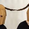 two figures wearing cardboard helmets with their pony tails tied together