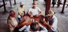 five men sit around a wooden table playing dominos